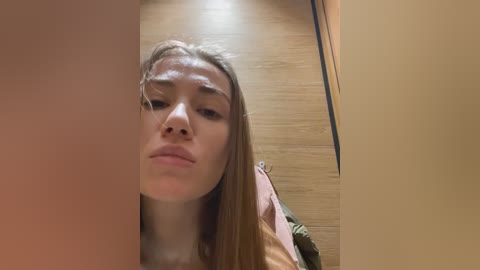 Media: A video of a young woman with light skin, straight brown hair, and minimal makeup, standing in a wooden hallway with a mirror reflection. She has a neutral expression, looking slightly to the side.