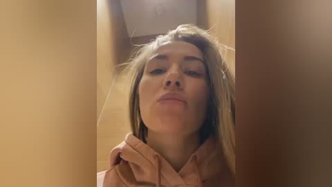 Media: A close-up video of a young woman with light skin and straight, shoulder-length blonde hair, wearing a pink hoodie, taken indoors, showing a narrow hallway with beige walls.