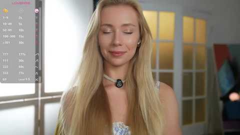 Media: Video of a serene blonde woman with long hair, wearing a white lace top, standing indoors near a window with a calendar app displayed on a screen.