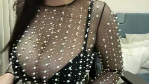 Media: Video of a woman in a sheer black dress with pearl-like sequins, revealing her large breasts and cleavage. Background shows a messy bed and white curtains in a dimly lit room.