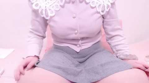 Media: Video of a woman sitting, wearing a white lace-trimmed blouse, gray skirt, and white wristbands, hands resting on knees, in a pastel-toned room.