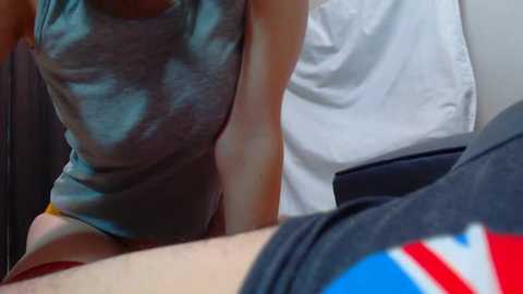 Media: Video of a woman in a grey tank top and orange shorts, leaning over a man in a blue shirt with a Union Jack design. Background shows a white pillow and dark wooden panel.