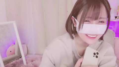 Media: Video of an Asian woman with a short bob haircut, wearing a white face mask, gray sweater, and holding a phone, smiling, in a cozy, dimly-lit room with a white wall and a plush rug.