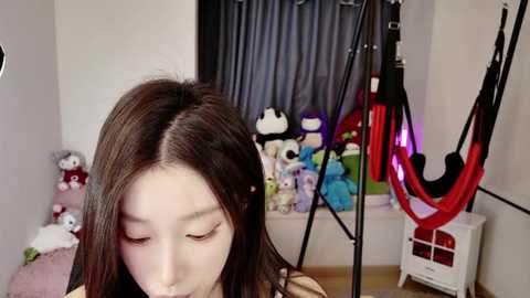 Media: Video of a young Asian woman with straight black hair, wearing a black top, sitting in a room with plush toys, a black curtain, and a red and black exercise ball.