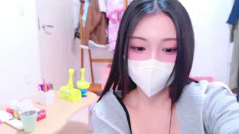 Media: Video of an Asian woman with long black hair, wearing a white face mask, a gray hoodie, and a pink necklace, sitting at a cluttered wooden desk in a brightly lit room with pink and yellow toys and a white door in the background.