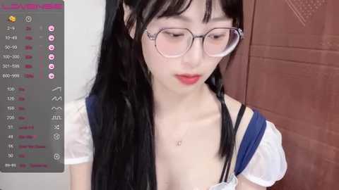 Media: Video of a young Asian woman with long black hair, wearing glasses, a blue and white dress, and a necklace, captured from the waist up, indoors.