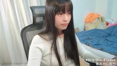 Media: Video of a young Asian woman with long, straight black hair and fair skin, wearing a white sweater, sitting on a black mesh chair. The room has a blue bedspread and white curtains.