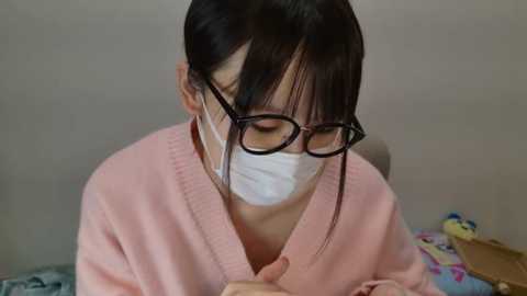 Media: Video of an Asian woman with straight black hair, wearing glasses, a pink V-neck sweater, and a white face mask, sitting indoors, holding her chin, looking down.