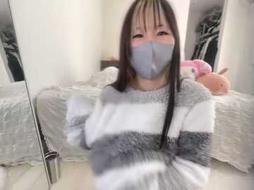 Media: Video of an Asian woman with long black hair, wearing a gray mask and gray and white striped sweater, sitting on a bed with plush toys, in a cozy, cluttered bedroom.