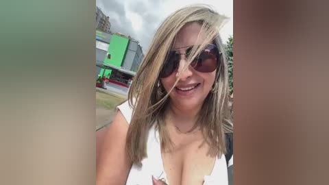 Media: Video of a smiling, blonde woman with sunglasses, wearing a white top, standing outdoors against a green building.