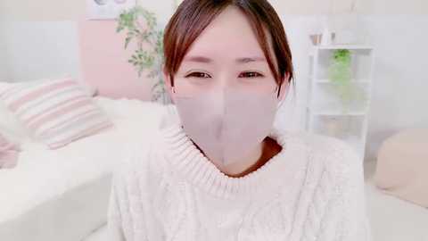 Media: Video of an Asian woman with light skin and short brown hair, wearing a white knit sweater and a surgical mask, in a pastel-toned bedroom with a bed, potted plants, and a white shelf in the background.