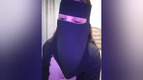 Media: Video of a person wearing a black niqab, covering their face except for their eyes, in a dimly lit room with a beige wall and a white curtain in the background.