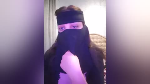 Media: A video of a woman with light skin, wearing a black mask and headband, stands in a dimly lit room with a beige couch and soft lighting.