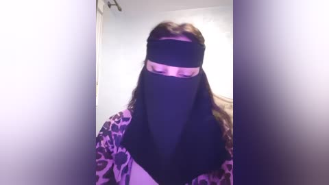 Media: Video of a person in a black hijab with a pink headband, wearing a purple floral robe. The background is a blurred, light-colored room with a doorway.