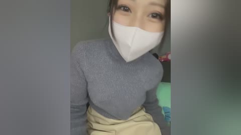 Media: A video of an Asian woman with a slender build, wearing a gray ribbed sweater and beige skirt, with a white surgical mask covering her mouth and nose.