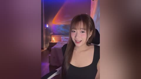 Media: Video of a smiling Asian woman with long brown hair, wearing a black sleeveless top, standing in a dimly lit bedroom with a bed, nightstand, and colorful wall art.