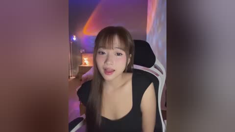 Media: Video of an Asian woman with long brown hair, wearing a black top, seated in a gaming chair, lit by purple and orange ambient lighting in a dimly-lit room.