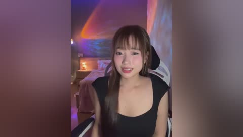 Media: Video of an Asian woman with long brown hair, wearing a black top, sitting in a dimly lit room with a colorful abstract painting and a chair behind her.