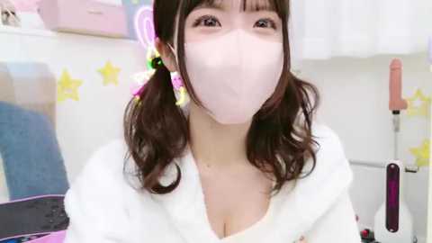 Media: A video of a young Asian woman with wavy brown hair, wearing a white mask, a white robe, and large earrings, standing in a brightly lit room with white walls and yellow star decals.