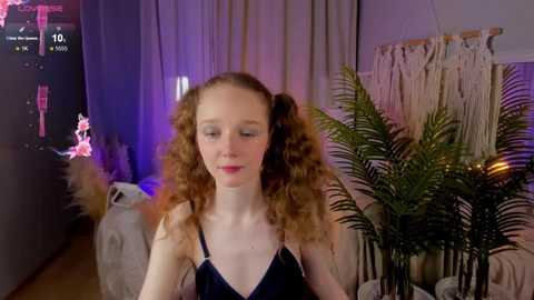 Media: Video of a young woman with curly red hair, wearing a dark blue camisole, standing in a cozy, dimly lit room with a white macram\u00e9 wall hanging, green ferns, and a lit candle.