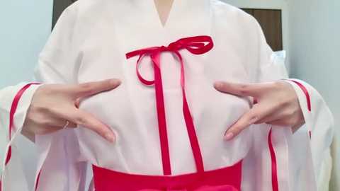 Media: Video of a woman wearing a white kimono with red trim, a red bow, and a red sash, hands resting on her chest. Background shows a light blue wall and a wooden door.