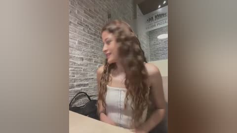 Media: Video of a fair-skinned woman with long, wavy brown hair, wearing a white spaghetti-strap top, sitting at a counter in a modern, gray brick-walled cafe.