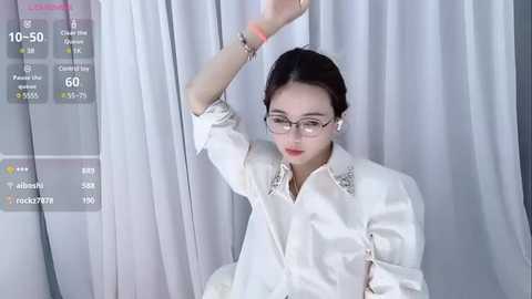 Media: Video of an East Asian woman with glasses, fair skin, and dark hair in a white blouse with sequin details, standing in front of a white curtain, holding a microphone.