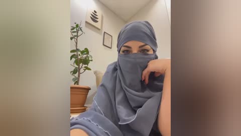 Media: Video of a woman in a gray hijab, sitting in a beige room with a potted plant and framed artwork on the wall.