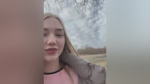 Media: Video of a young woman with long blonde hair, light skin, and pink lips, wearing a light pink top and grey jacket, standing outdoors under cloudy sky with barren trees and distant fields in the background.