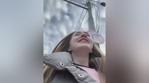 Media: A video shows a laughing young woman with long brown hair, wearing a pink shirt and gray puffy jacket, holding a pink and white traffic sign. The background is a cloudy sky.