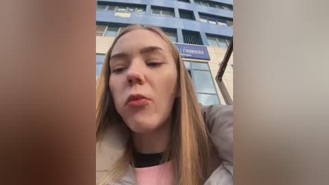 Media: A video of a young Caucasian woman with long blonde hair, fair skin, and closed eyes, wearing a pink sweater and black choker, standing in front of a blue and white building.