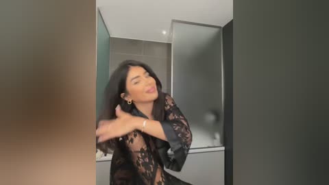 Media: Video of a woman with long, dark hair, wearing a sheer black lace robe, smiling and dancing in a modern bathroom with glass shower and tiled walls.