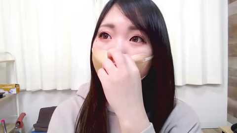 Media: Video of an East Asian woman with long black hair, light skin, and wide eyes, gagged with tape, sitting indoors with white curtains and scattered makeup brushes in the background.