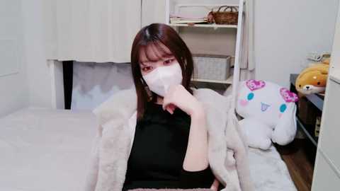 Media: A video of an East Asian woman with straight, shoulder-length dark brown hair, wearing a white mask, black top, and a light jacket, sitting on a white bed in a minimalist room with a stuffed toy and shelves.