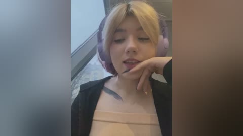 Media: Video of a young Asian woman with blonde hair and purple headphones, wearing a beige top and black jacket, sitting indoors, slightly blurry background.