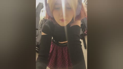 Media: Video of a person with a short, pink wig, wearing a black off-the-shoulder top, red plaid skirt, and black arm sleeves, posing in a dimly lit room with clothes and a mirror in the background.