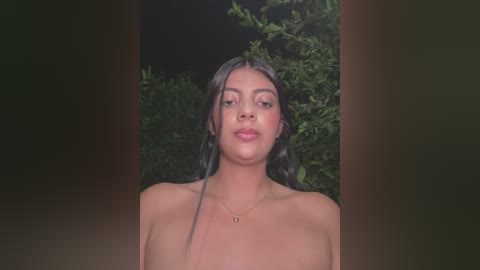 Media: Video of a young woman with wet, dark hair and medium brown skin, standing topless outdoors against a backdrop of dark foliage.