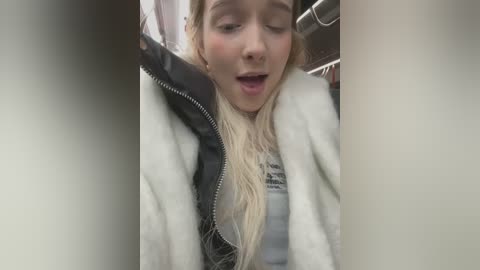 Media: Video of a blonde woman with fair skin, wearing a black jacket over a white shirt, leaning against a metal railing, mouth open in a surprised expression.