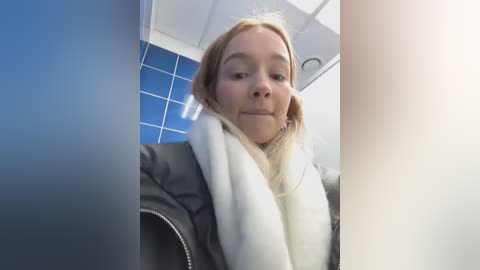 Media: Video of a blonde woman in a public restroom with blue tiled walls and white ceiling. She wears a black leather jacket and white scarf, looking slightly to the right.