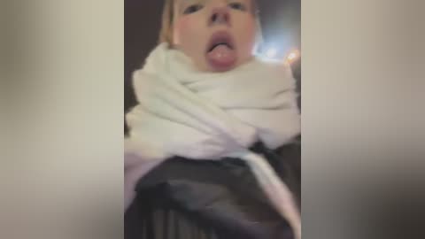 Media: A blurry video captures a person with a shocked or surprised expression, tongue out, wrapped in a white blanket. The background is dark and out of focus, emphasizing the subject.