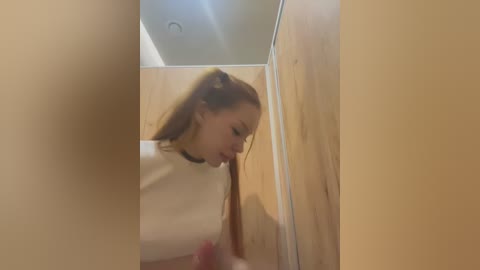 Media: Video of a young woman with long, red hair in a ponytail, wearing a black choker, standing in a wooden-paneled bathroom with a mirror. Her reflection is blurred, creating a surreal, distorted effect.