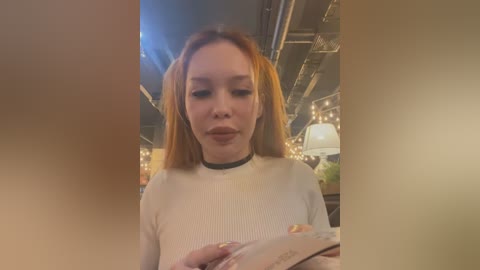 Media: Video of a young woman with straight, light brown hair, wearing a white ribbed sweater and black choker, reading a book in a cozy, dimly lit caf\u00e9 with string lights and exposed pipes.