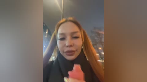 Media: A video of an Asian woman with long, straight, light brown hair, wearing a black coat and a pink scarf, smiling, with a blurred urban cityscape in the background.