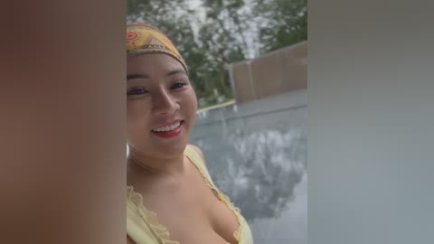 Media: Video of a smiling Asian woman with medium skin tone, wearing a yellow headscarf, partially unbuttoned yellow blouse, and red lipstick, standing near a pool with blurred greenery in the background.