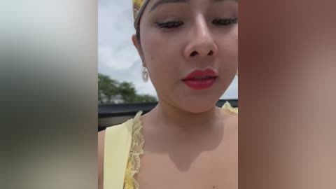 Media: Video of an Asian woman with light skin, red lipstick, and yellow headband, wearing a yellow blouse, looking downward, with blurred outdoor background.