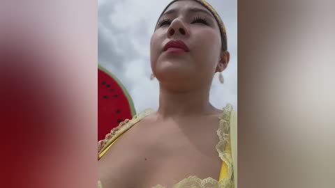 Media: Video of an Asian woman with light skin, wearing yellow lace top, head tilted back, holding a red watermelon, smiling. Background shows cloudy sky.