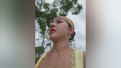 Media: Video of an Asian woman with light skin, wearing a yellow headscarf and a lace-trimmed yellow top, gazing upwards against a blurred green tree background.