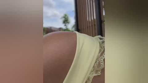 Media: Video of a close-up of a woman's bare buttocks, clad in a lacy, light yellow thong, with a blurry outdoor background featuring trees and a blue sky.