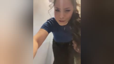 Media: A blurry video of a woman with fair skin and wavy brown hair, wearing a blue top and black pants, seen from behind, possibly in a bathroom.