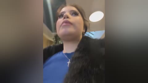 Media: Video of a young woman with fair skin, brown hair, and dark eyeliner, wearing a blue shirt and black fur-lined coat, standing in an indoor setting with a blurred background.